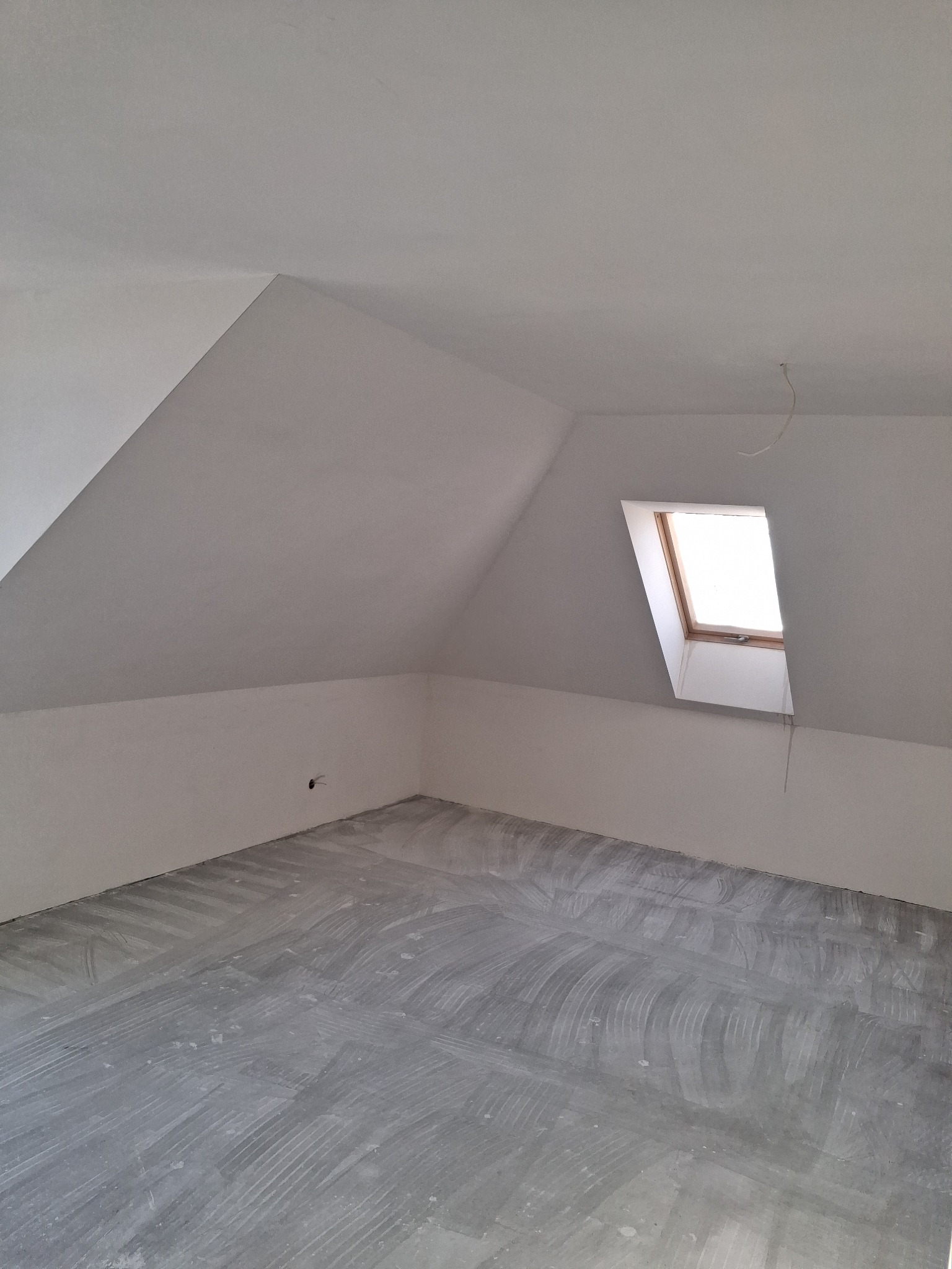 Poddasze ze skośnym dachem i oknem dachowym po szpachlowaniu. Widoczne ślady po szpachli na podłodze i ścianach. Przygotowanie do dalszych prac wykończeniowych.