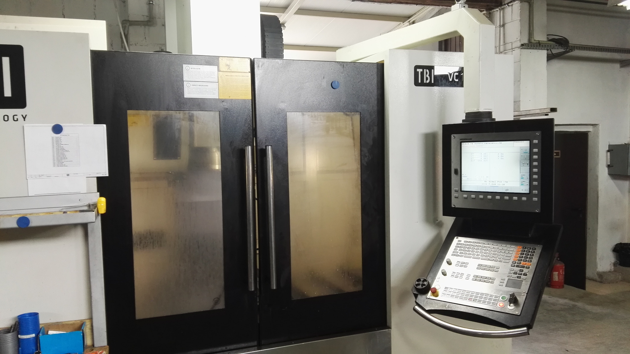 Zamknięta frezarka CNC z panelem sterowania Heidenhain i instrukcją obsługi przyczepioną magnesem.
