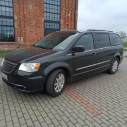 Ciemny minivan Chrysler Town & Country zaparkowany na brukowanym podjeździe przed ceglanym budynkiem z dużymi oknami.