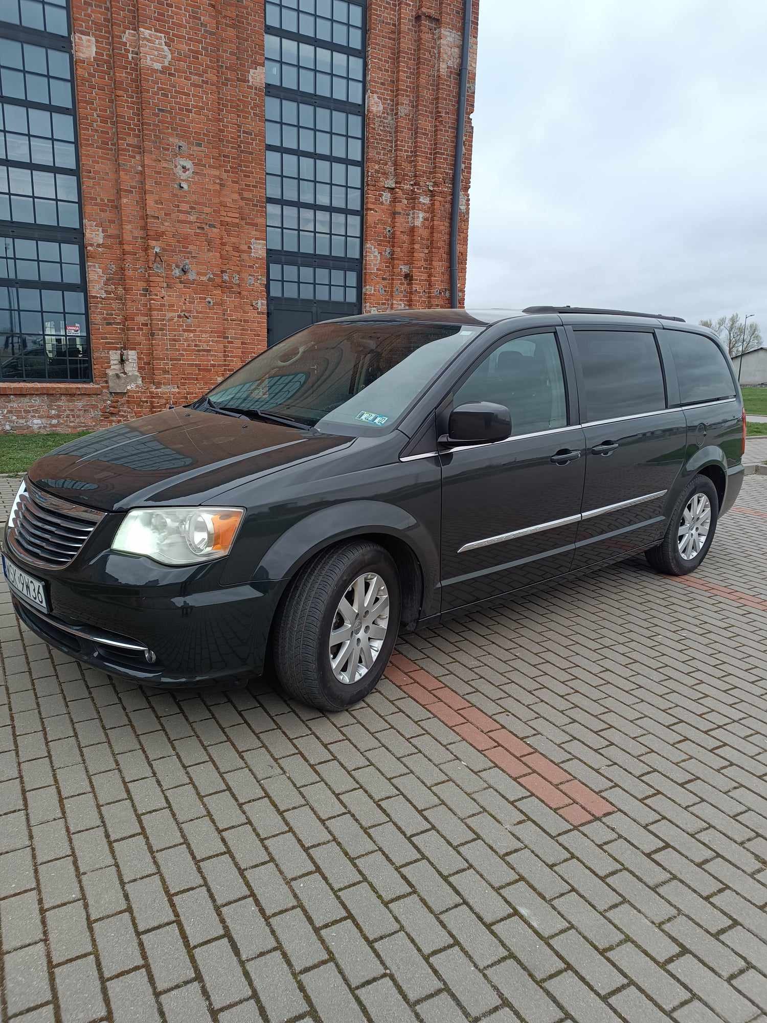 Ciemny minivan Chrysler Town & Country zaparkowany na brukowanym podjeździe przed ceglanym budynkiem z dużymi oknami.