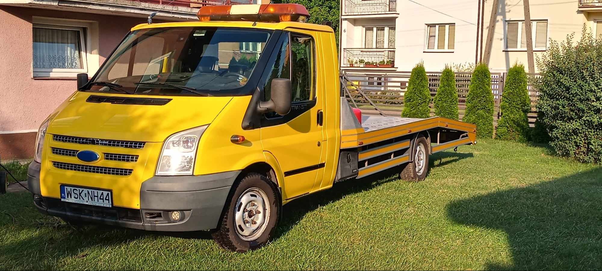 Żółta laweta Ford Transit na trawniku przed budynkiem mieszkalnym, widok z boku na przód pojazdu, z widoczną tablicą rejestracyjną.