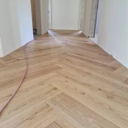 Ułożona parkietem w jodełkę jasna podłoga w nowym mieszkaniu, widoczny fragment różowego węża wzdłuż ściany.