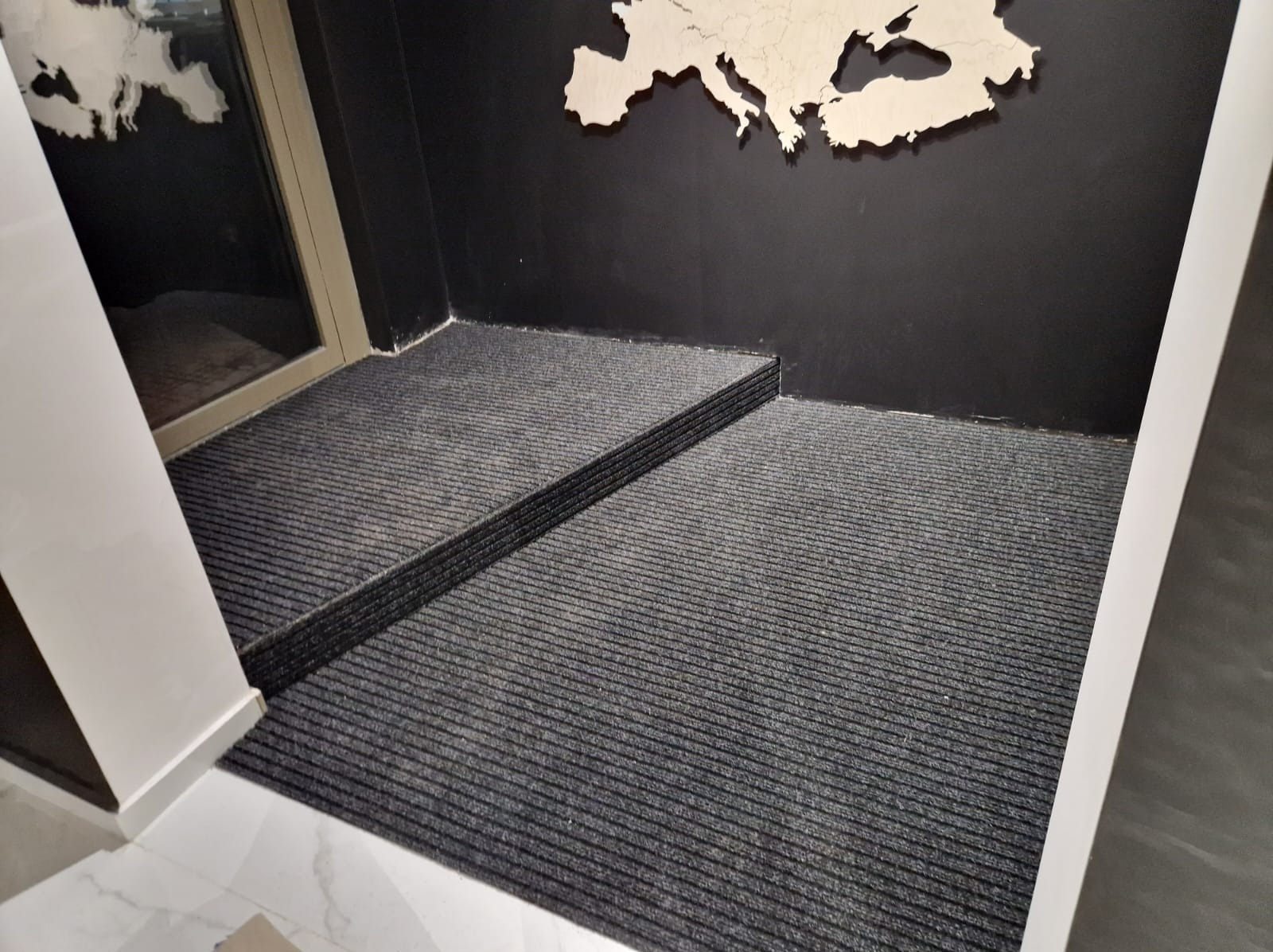 Szara, prążkowana wycieraczka wpuszczona w podłogę przy wejściu, z widoczną drewnianą mapą Europy na czarnej ścianie.