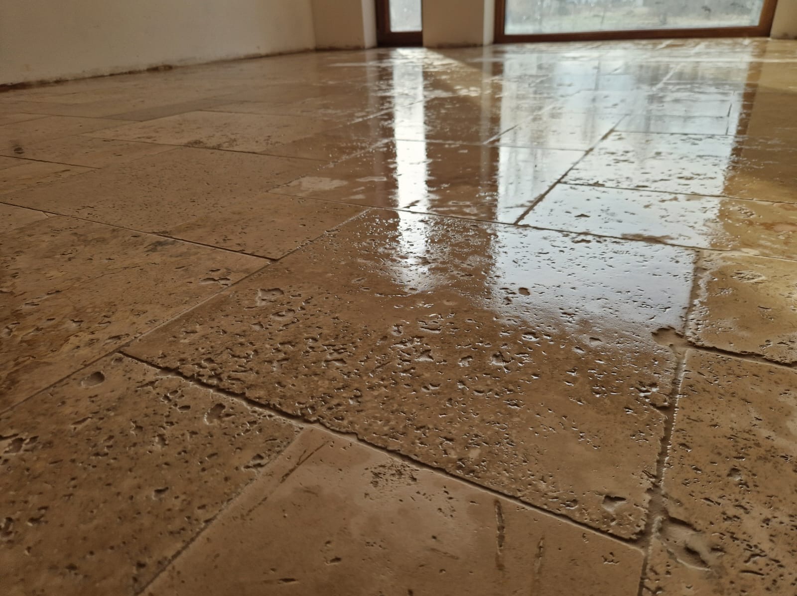 Świeżo ułożona, mokra podłoga z płytek trawertynowych, odbijająca światło okien w pomieszczeniu. Widoczne nierówności i porowata struktura kamienia.