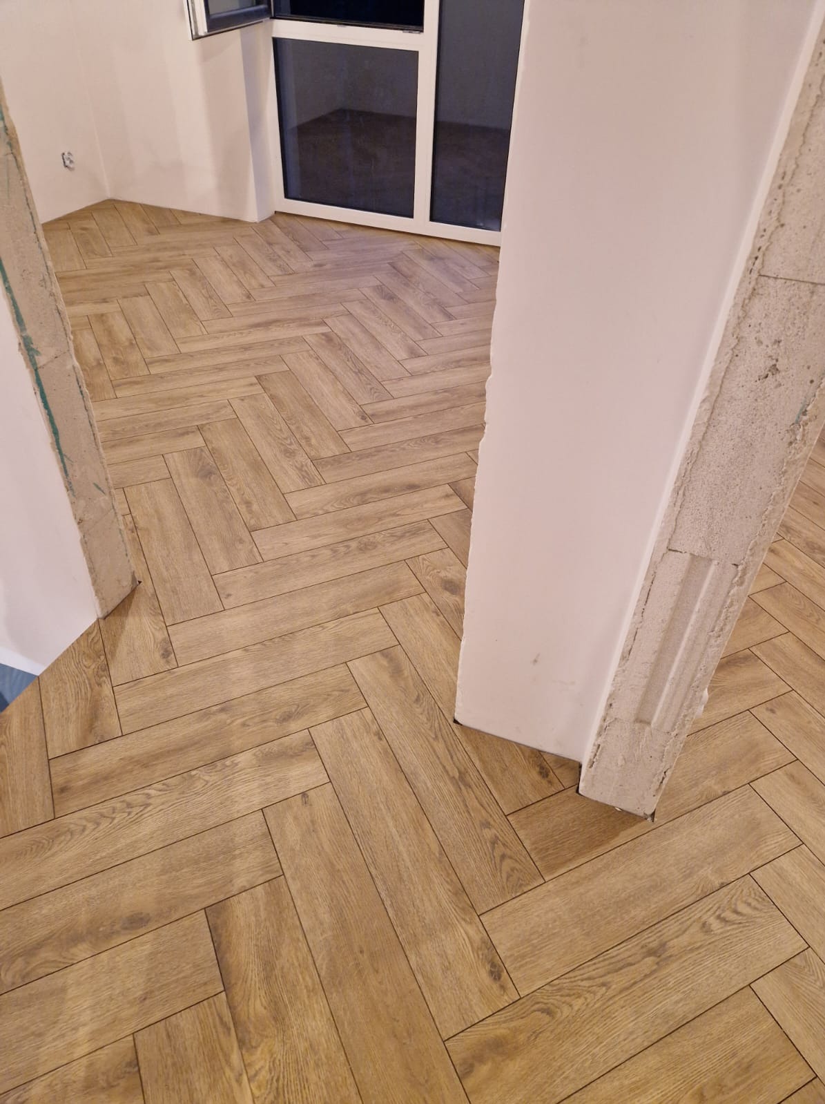 Ułożona w jodełkę podłoga z paneli imitujących drewno, widok z progu, surowy betonowy filar po lewej stronie, białe ściany i okno w tle.