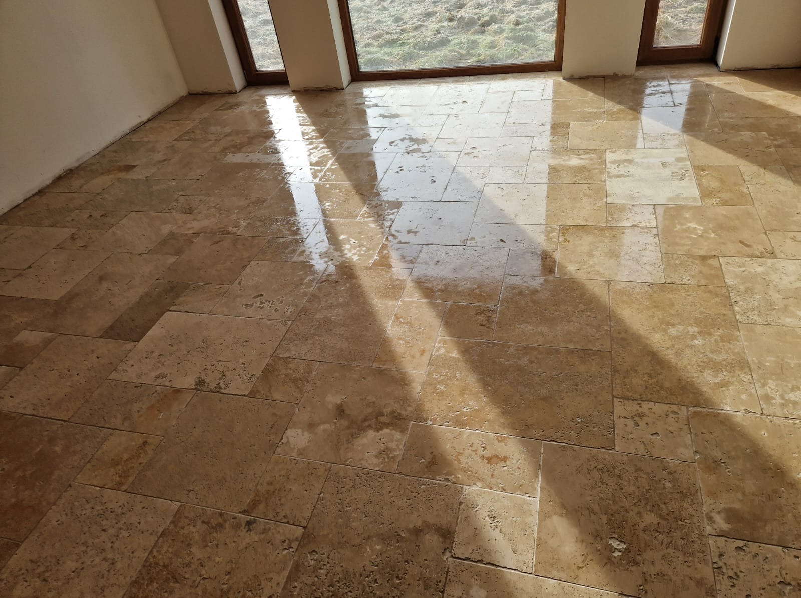 Wyłożona trawertynem podłoga w jasnym pomieszczeniu, mokra powierzchnia odbija światło słoneczne wpadające przez okno.