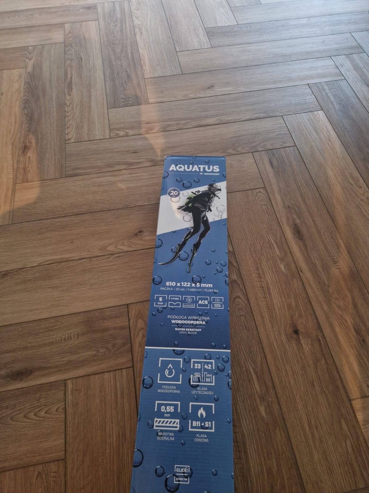 Opakowanie paneli winylowych Aquatus wodoodpornych leżące na podłodze z paneli ułożonych w jodełkę. Na opakowaniu grafika nurka.