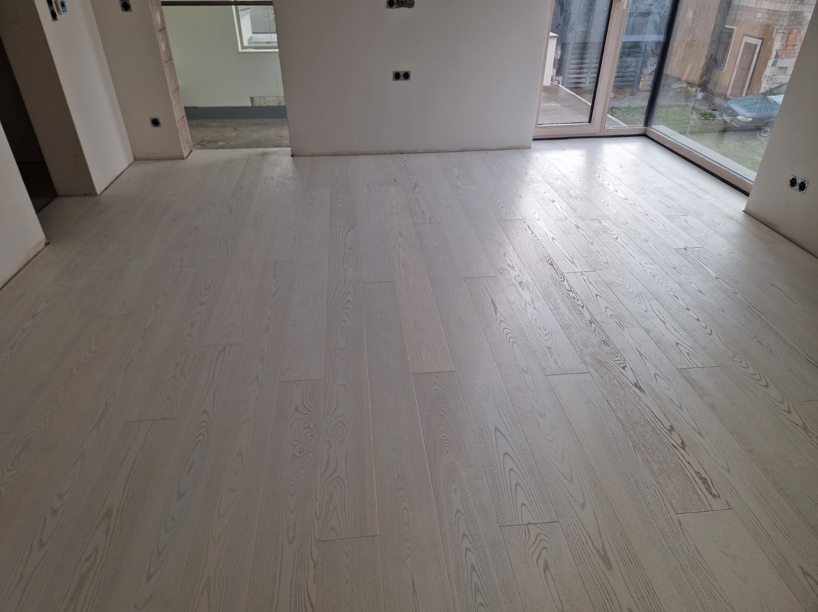 Jasna, drewniana podłoga ułożona w nowym wnętrzu z dużym oknem. Widoczne gniazdka elektryczne na ścianach, surowy stan wykończenia.