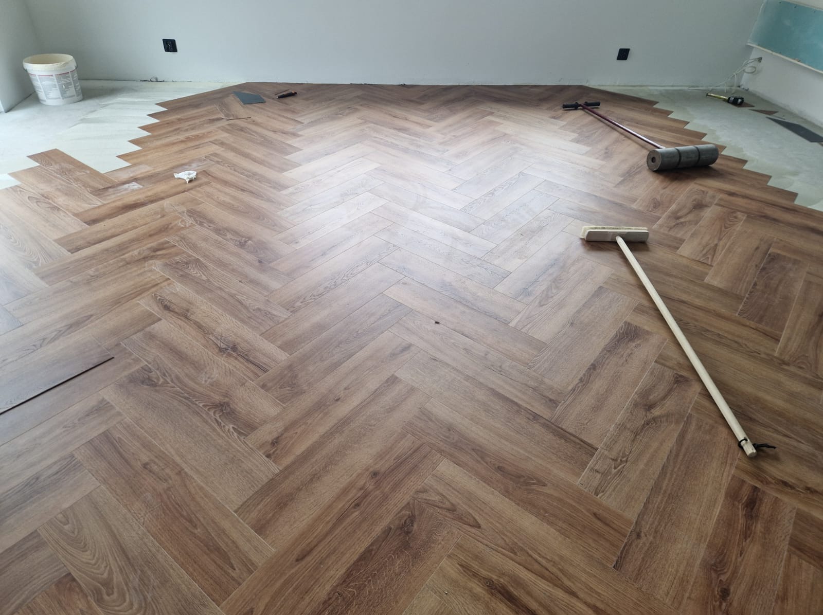 Układanie paneli podłogowych w jodełkę, widoczne narzędzia: wałek dociskowy i szczotka, częściowo ułożona podłoga.