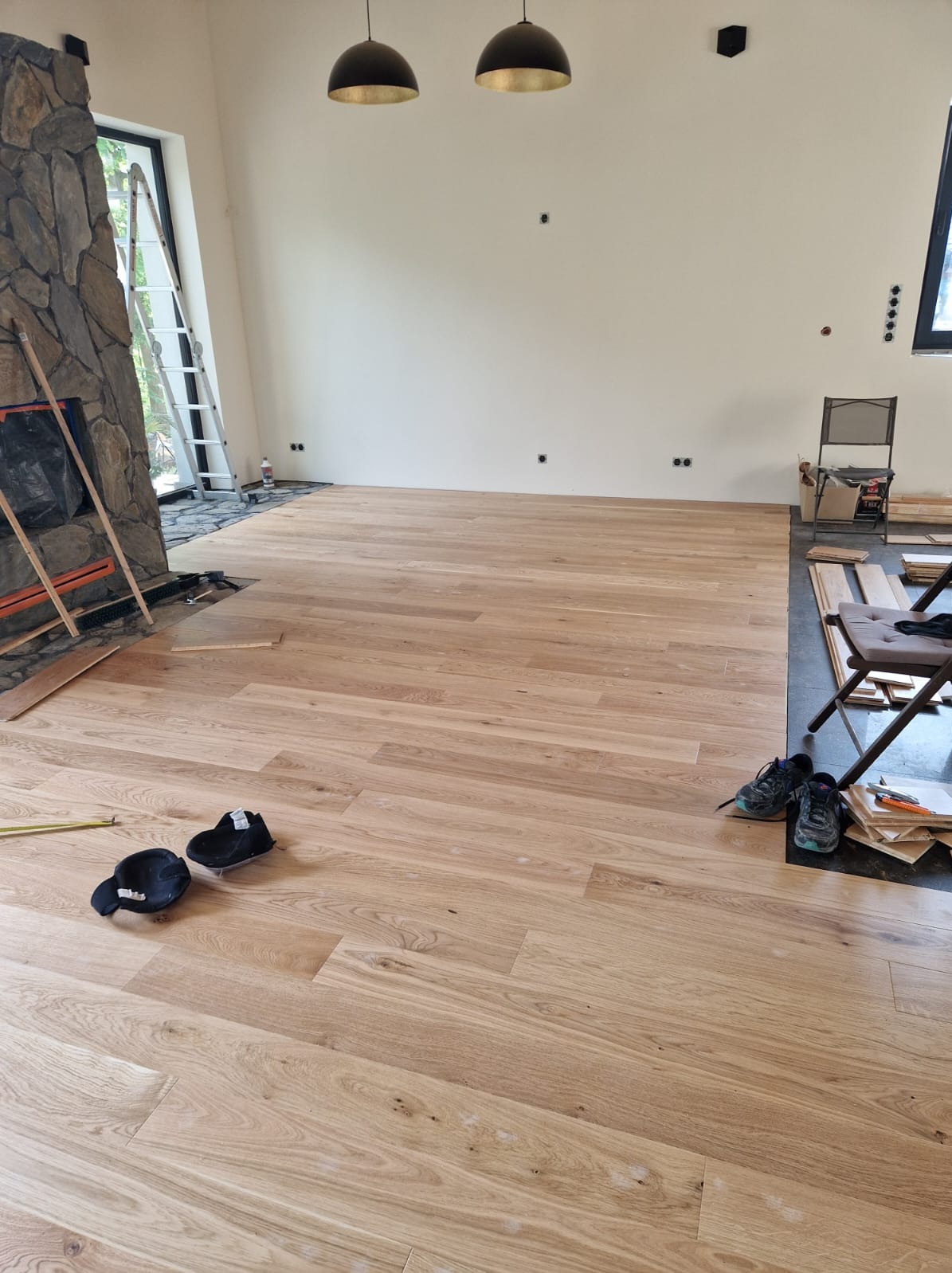 Układanie jasnej, drewnianej podłogi w przestronnym salonie z kamiennym kominkiem, widoczne narzędzia i obuwie robocze, surowe ściany i wiszące lampy.