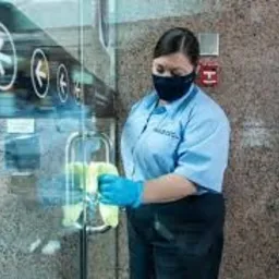 Pracownica w uniformie i masce ochronnej czyści szklane drzwi wejściowe żółtą ściereczką, nosząc niebieskie rękawice ochronne. Na drzwiach widoczne symbole strzałek kierunkowych.