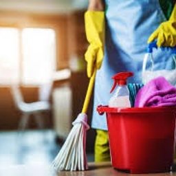 NEXUS ENTERPRISE SPÓŁKA Z OGRANICZONĄ ODPOWIEDZIALNOŚCIĄ - Czerwone wiadro z płynami do czyszczenia, spryskiwaczem i ściereczkami z mikrofibry, obok mop z żółtymi rękawicami na tle rozmytego wnętrza mieszkania z oknem.