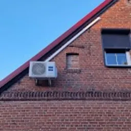 Zewnętrzna jednostka klimatyzacji Haier zamontowana na ceglanej ścianie szczytowej domu pod okapem dachu, widok z dołu na tle błękitnego nieba.