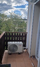 Kompaktowa jednostka klimatyzacyjna zewnętrzna, zamontowana na balkonie z brązową balustradą, widok na drzewa i budynki w tle, pochmurne niebo.