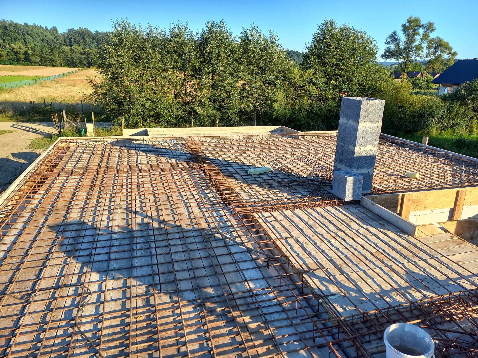 Zbrojenie stropu budynku żelbetowego przygotowane do zalania betonem, widoczne szalunki i komin wentylacyjny z bloczków betonowych, w tle drzewa i zabudowa jednorodzinna.