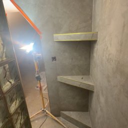 True COLORS - Wykończona wnęka prysznicowa z półkami z płytek w odcieniach szarości, oświetlona lampą budowlaną. Widoczny fragment szklanej ścianki działowej.