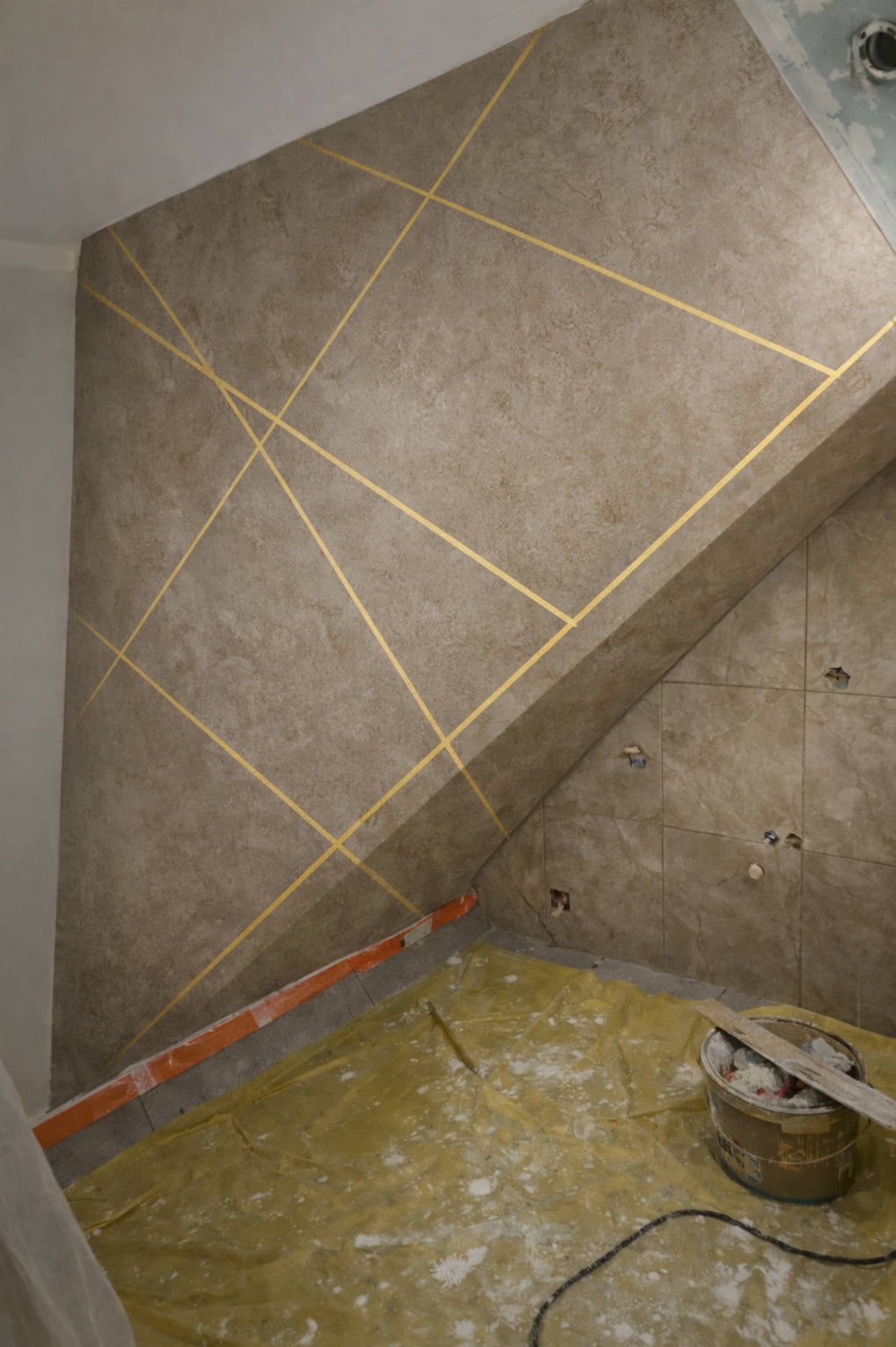 Ściana z dekoracyjną, złotą geometrią w trakcie remontu łazienki na poddaszu. Podłoga zabezpieczona folią, widoczne narzędzia i wiadro z zaprawą.