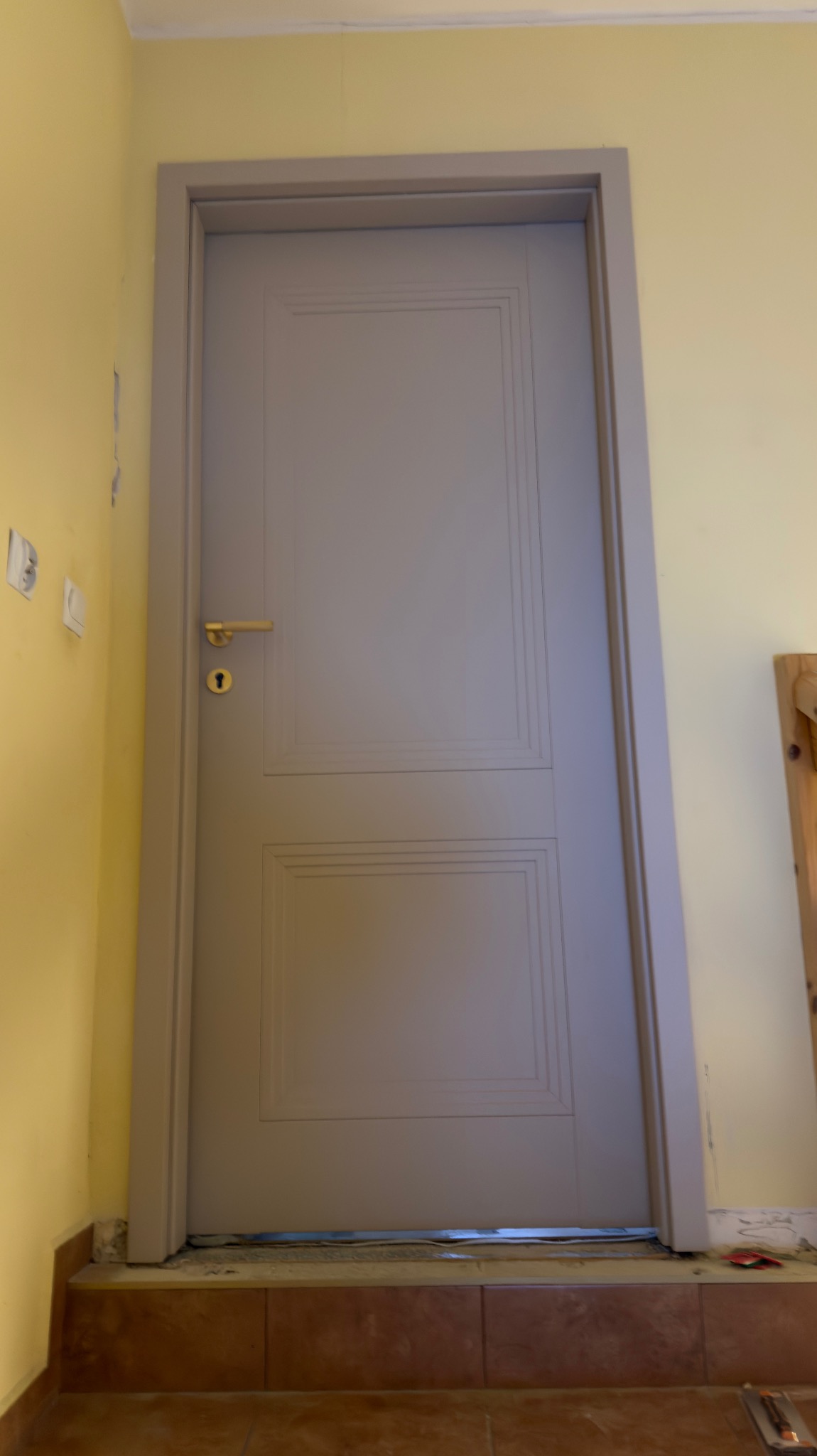 Szare, dwuskrzydłowe drzwi wewnętrzne z złotą klamką, osadzone w żółtej ścianie. Widoczne ślady prac wykończeniowych przy podłodze.