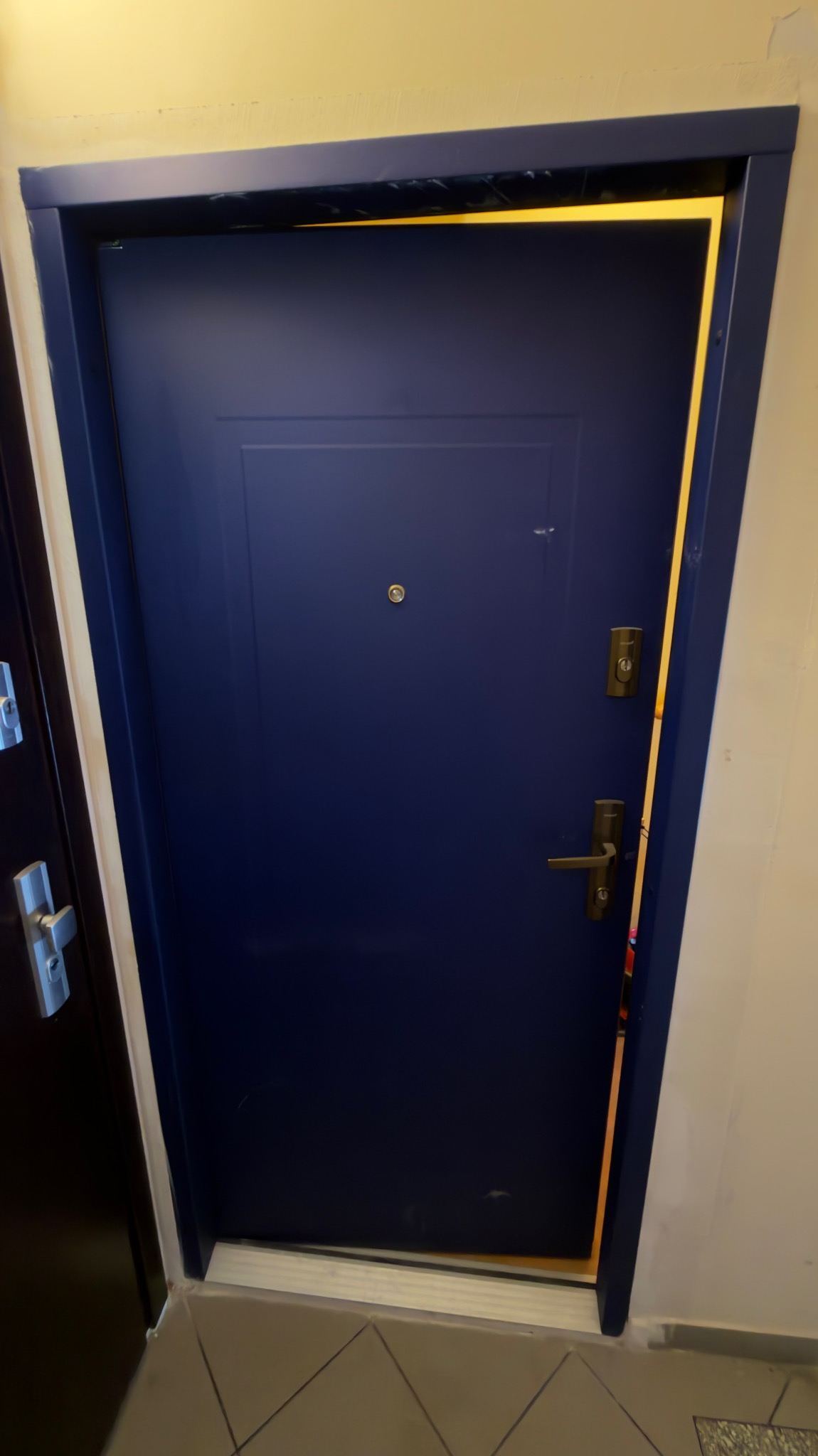 Niebieskie, gładkie drzwi wejściowe z wizjerem i srebrną klamką, osadzone w ciemnej futrynie. Widoczny fragment podłogi z szarych płytek.