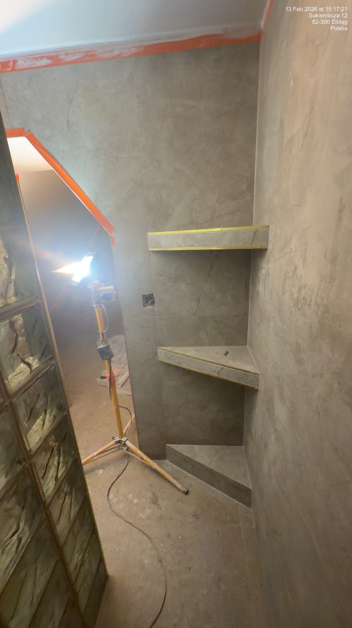 Wykończona wnęka prysznicowa z półkami z płytek w odcieniach szarości, oświetlona lampą budowlaną. Widoczny fragment szklanej ścianki działowej.