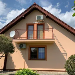 Jasnożółta elewacja domu jednorodzinnego z brązowym dachem, balkonem z pomarańczową balustradą, dwoma oknami z brązowymi ramami, dwoma klimatyzatorami AUX i anteną satelitarną TechniSat.