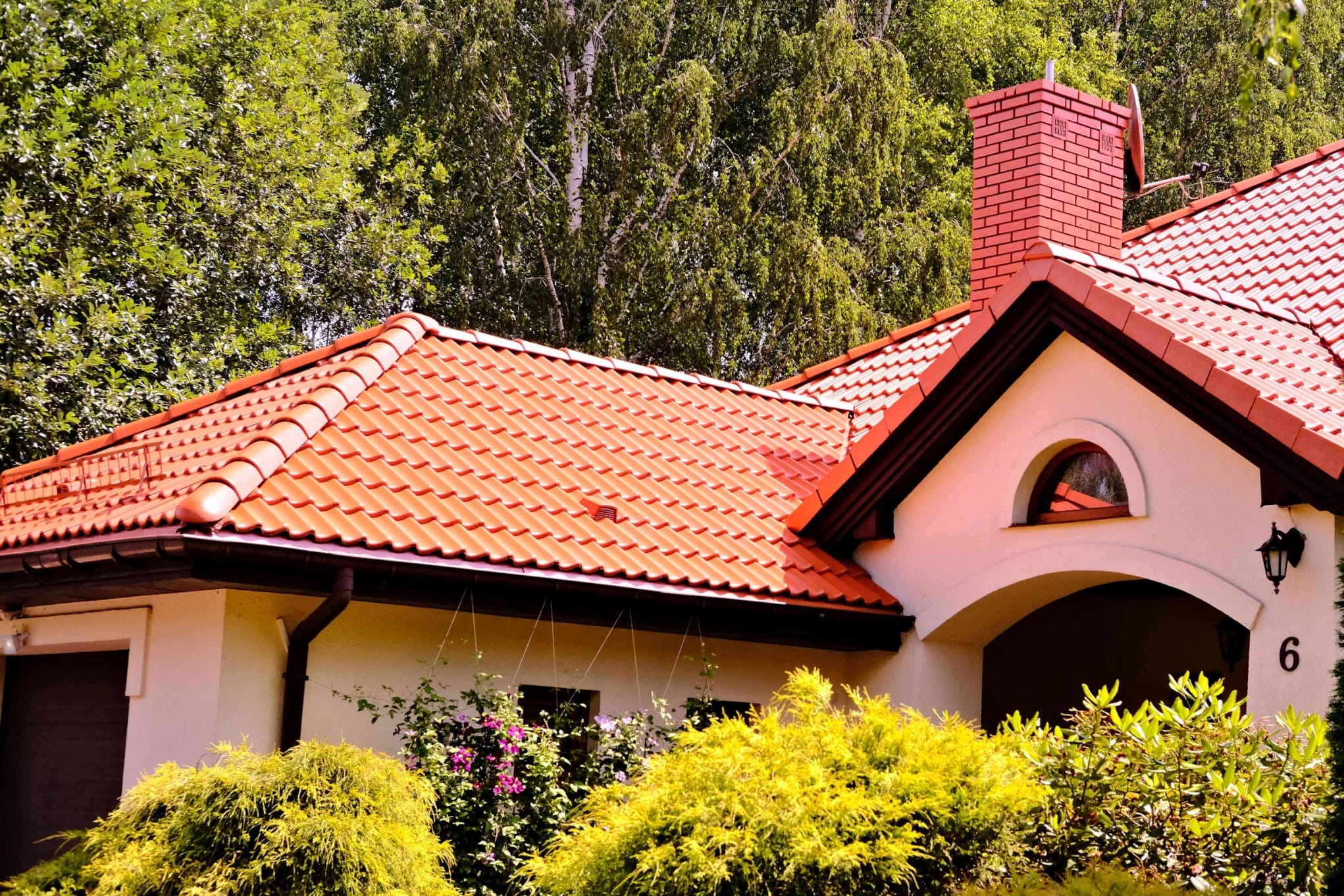 Dom jednorodzinny z jasną elewacją, czerwoną dachówką i ceglanym kominem, otoczony zielenią drzew i krzewów, numer domu '6' widoczny na ścianie.