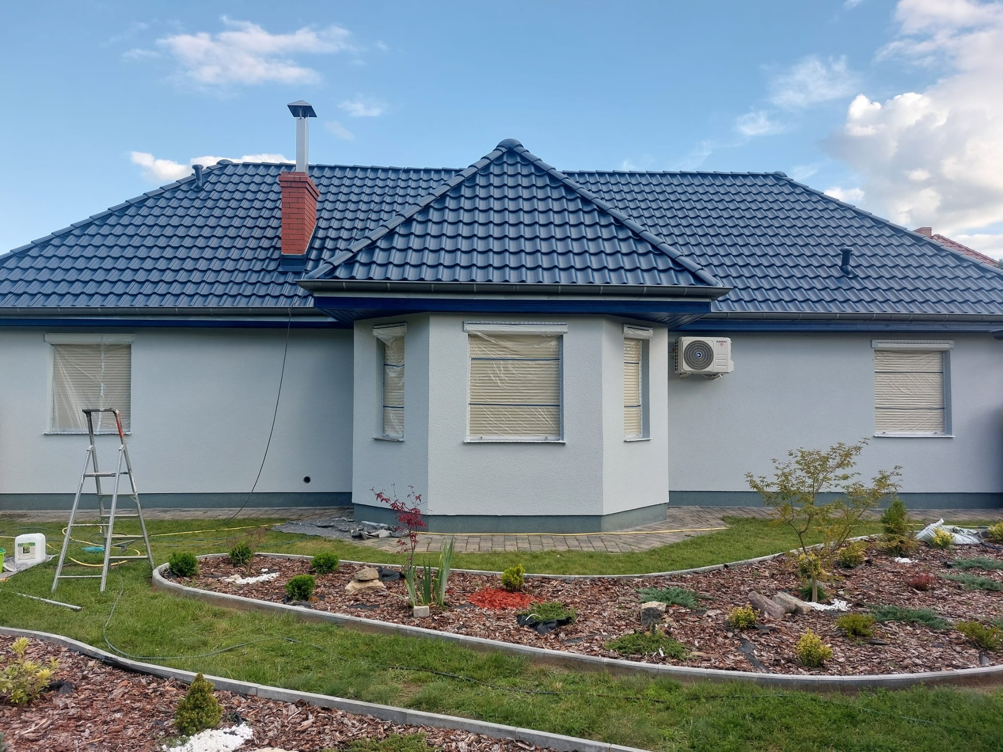 Dom jednorodzinny z szarą elewacją i ciemną dachówką, okna zabezpieczone folią, widoczna drabina i przygotowany teren wokół domu, sugerujące prace wykończeniowe.