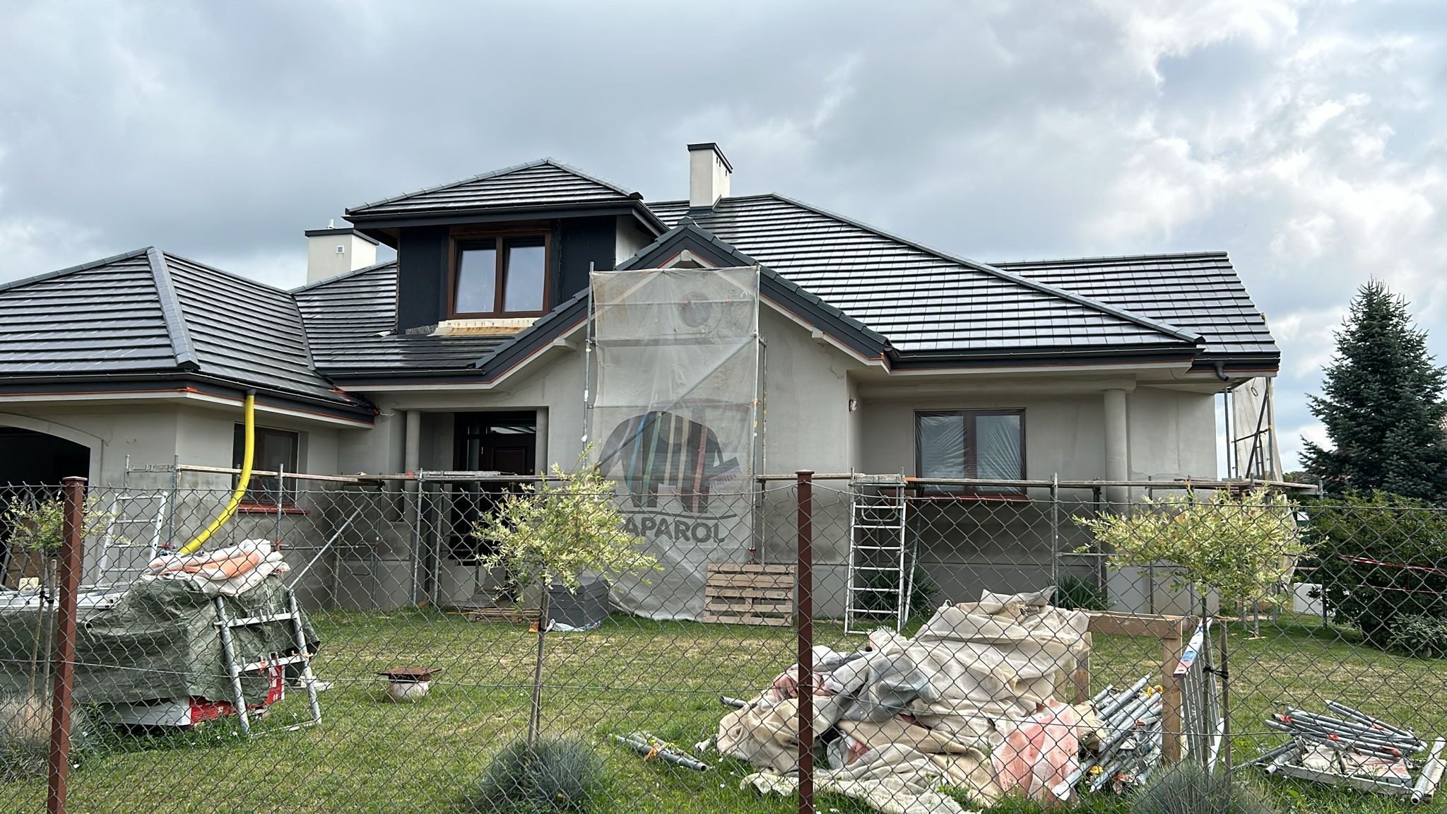 Dom jednorodzinny w trakcie prac elewacyjnych, widoczne rusztowania, siatka ochronna z logo producenta farb Caparol oraz materiały budowlane na trawniku.