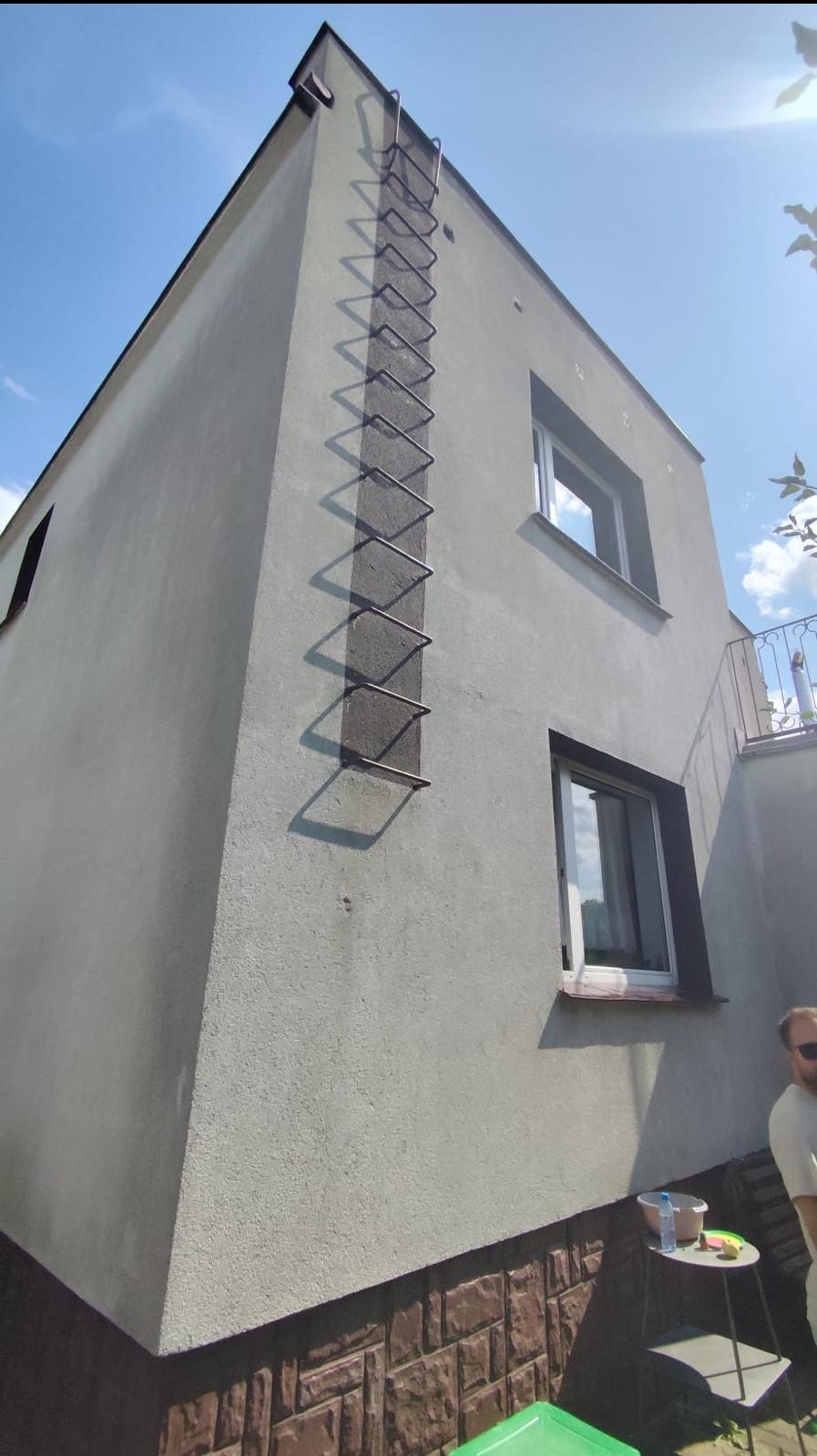 Szara elewacja dwupiętrowego domu z zamontowaną metalową drabinką przeciwpożarową, widoczne dwa okna z ciemnymi ramami, cokół wykończony brązową imitacją kamienia, fragment balkonu i niebieskie niebo.