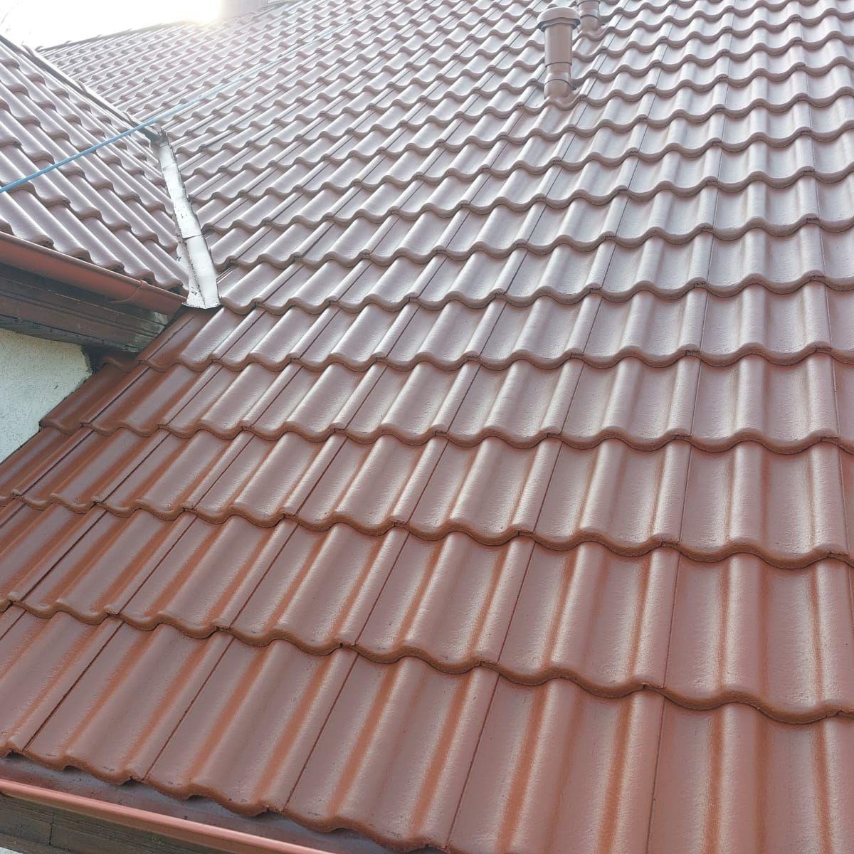 Czerwona, metalowa dachówka na skośnym dachu domu, widoczny system rynnowy i komin wentylacyjny