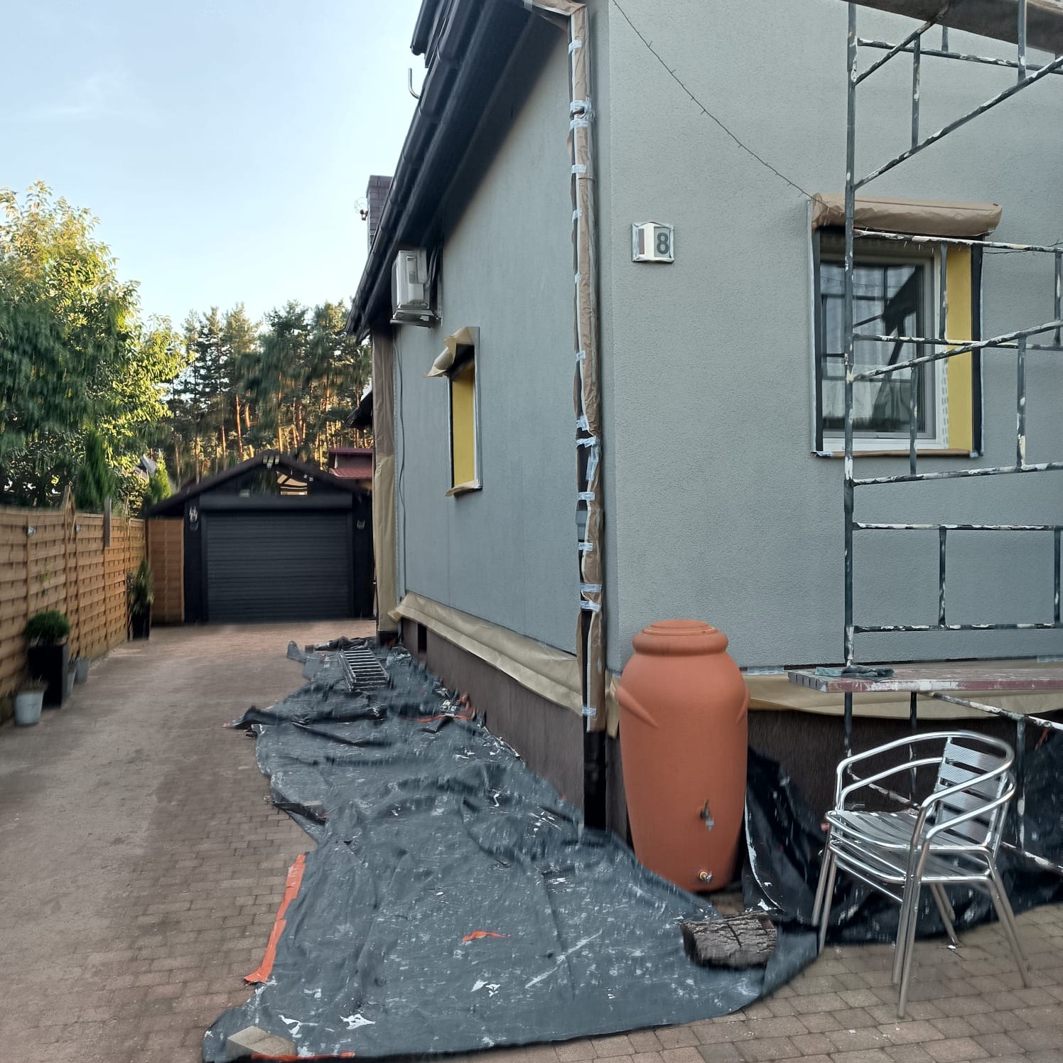 Dom jednorodzinny z szarą elewacją w trakcie prac wykończeniowych, widoczne rusztowanie, zabezpieczona folią podstawa budynku, zbiornik na deszczówkę i numer domu 18.