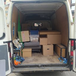 Auto pomoc, serwis, przeprowadzki - Łukasz Knap - Wnętrze białego busa wypełnione kartonami, skrzyniami i zabezpieczeniami do przewozu mebli i sprzętów podczas przeprowadzki, widoczne pasy transportowe i siatka zabezpieczająca, drewniana podłoga...