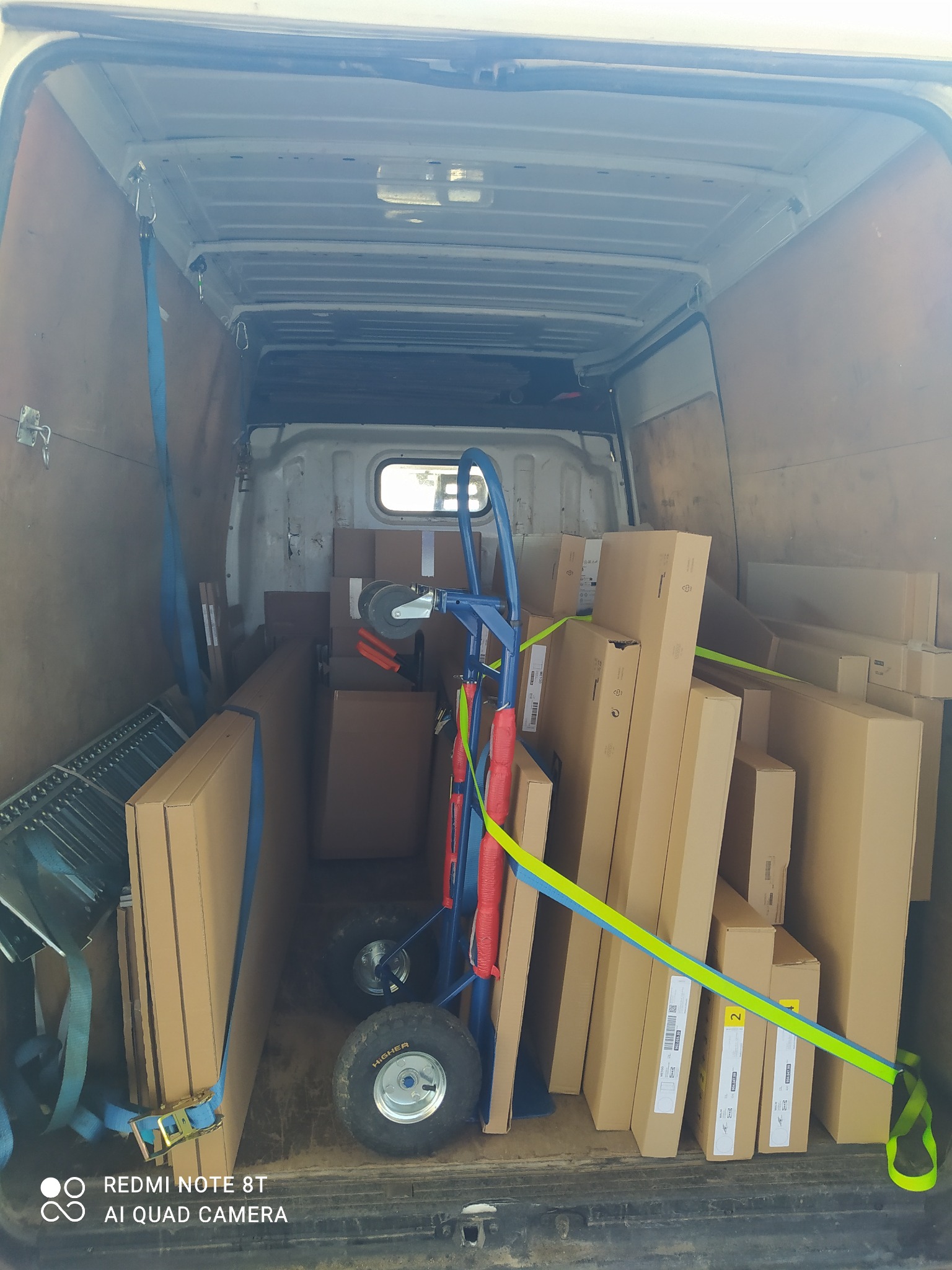 Wnętrze furgonetki załadowane kartonami, wózkiem transportowym z czerwonymi uchwytami i zabezpieczone niebieskimi pasami transportowymi.