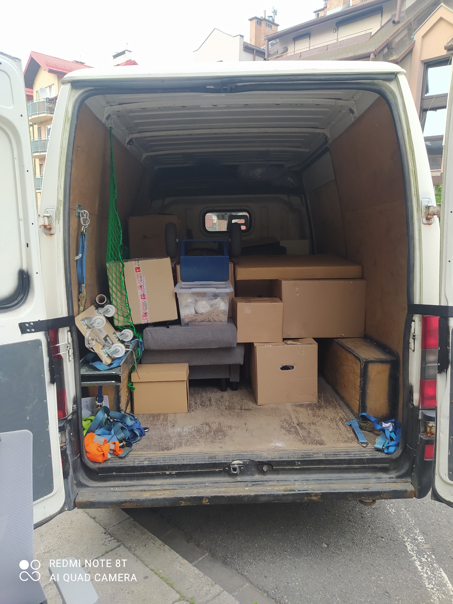 Wnętrze białego busa wypełnione kartonami, skrzyniami i zabezpieczeniami do przewozu mebli i sprzętów podczas przeprowadzki, widoczne pasy transportowe i siatka zabezpieczająca, drewniana podłoga...