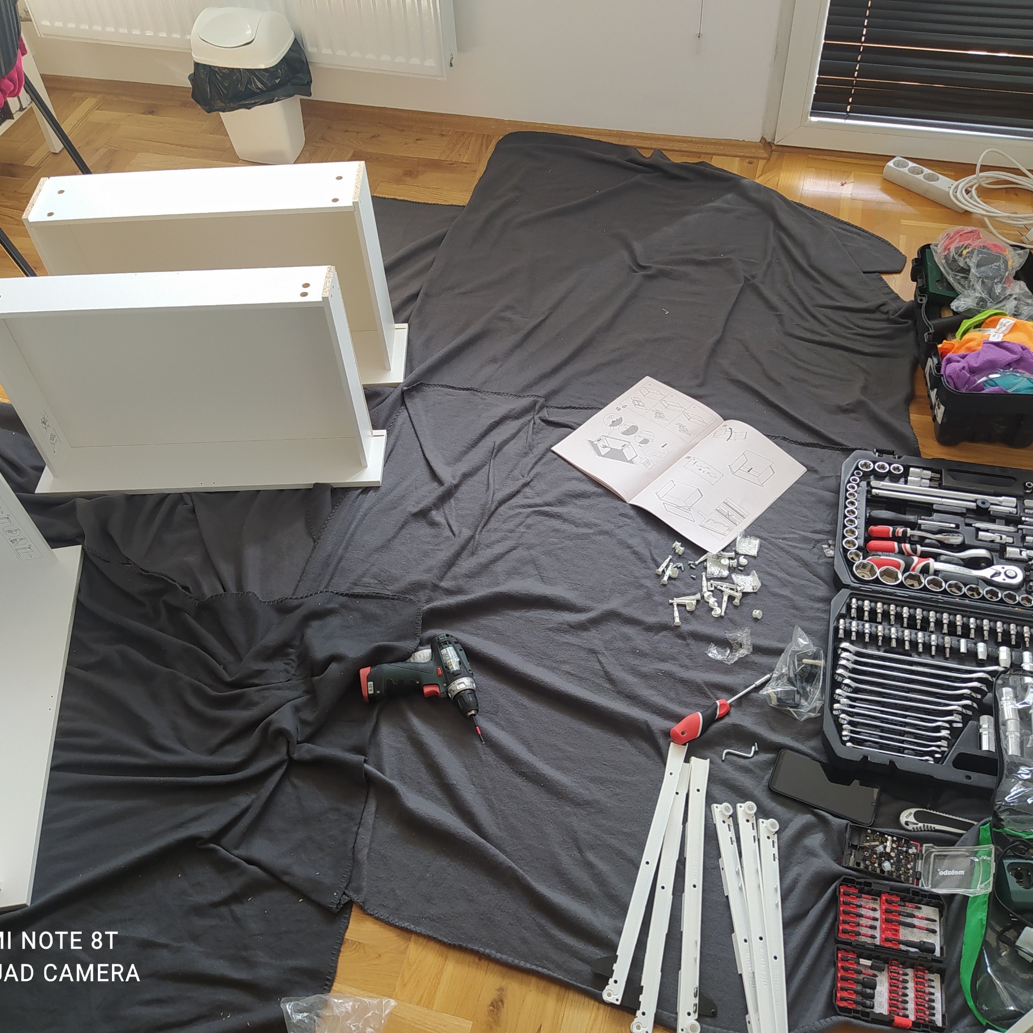 Rozłożone elementy białych szuflad na podłodze, instrukcja montażu, wkrętarka, zestaw narzędzi i akcesoriów do skręcania mebli na czarnej macie ochronnej.