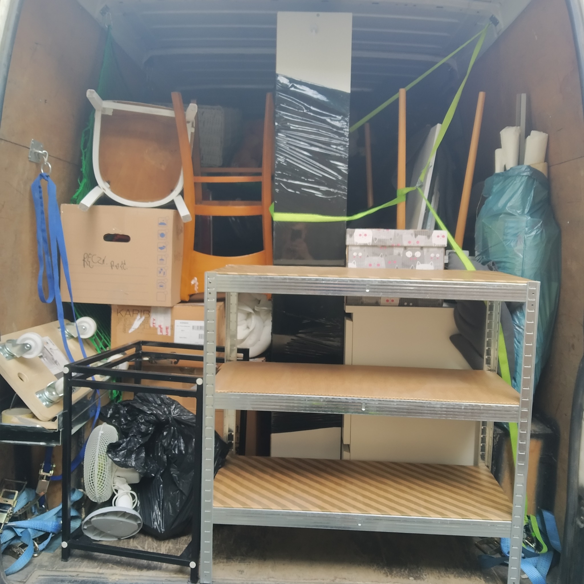 Wnętrze furgonetki załadowane meblami i kartonami zabezpieczonymi pasami, widoczny regał metalowy z drewnianymi półkami, krzesło z białymi nogami i karton z napisem 'Reczki. post.'.
