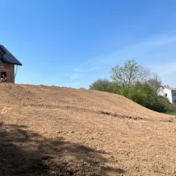 Mini koparka przy domu w budowie na wzgórzu z ziemi. Ślady opon na ziemi, błękitne niebo. Prace ziemne w toku, widok na okoliczne zabudowania i słupy energetyczne.