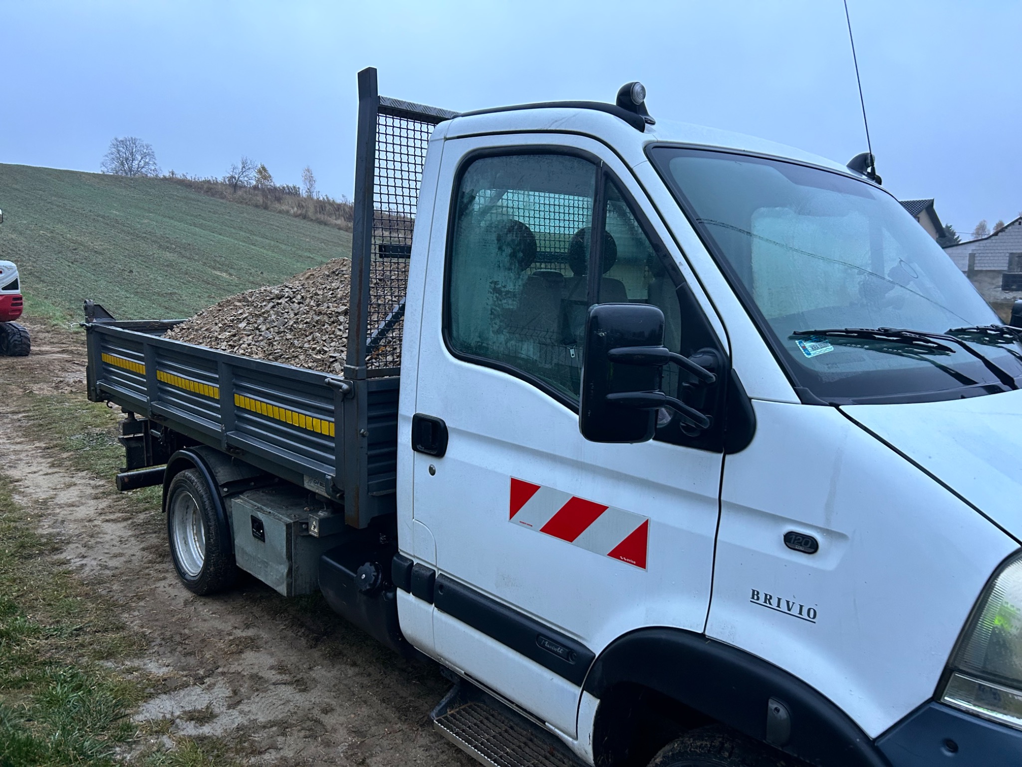 Ciężarówka wywrotka z pełną skrzynią gruzu lub kamieni na tle zielonego pola i pochmurnego nieba. Widoczny fragment małej koparki.