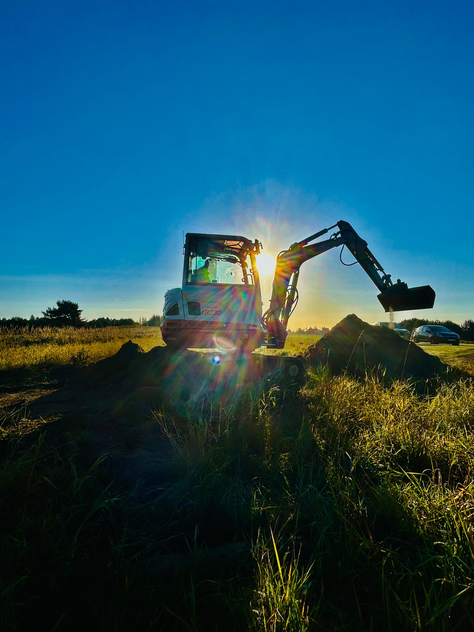 Mała koparka nabiera ziemię na tle wschodzącego słońca, z widoczną sylwetką operatora w kabinie i polną trawą w pierwszym planie.
