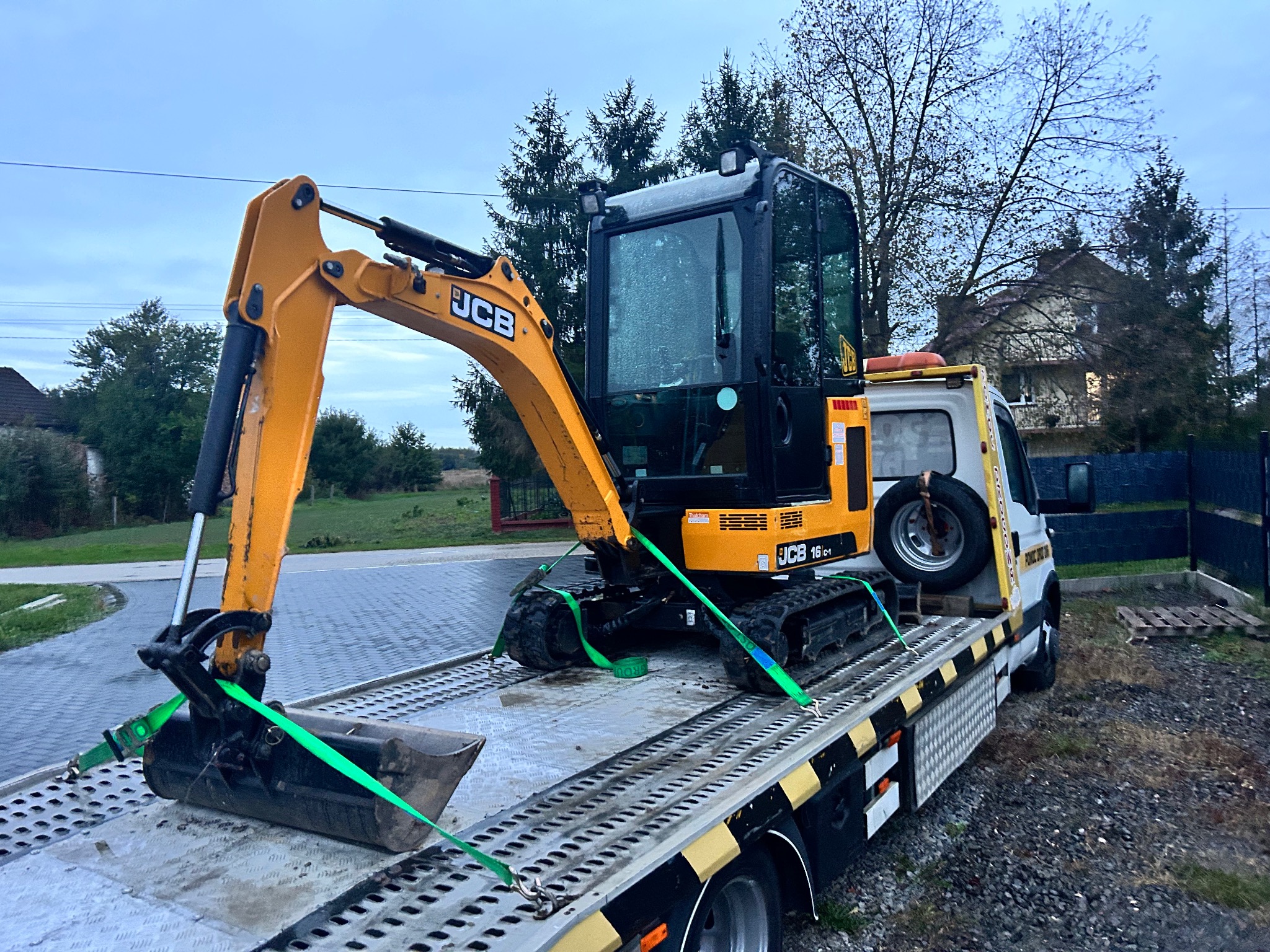 Żółta minikoparka JCB 16C-1 zabezpieczona zielonymi pasami transportowymi na platformie lawety, widok z boku, w tle dom i drzewa.