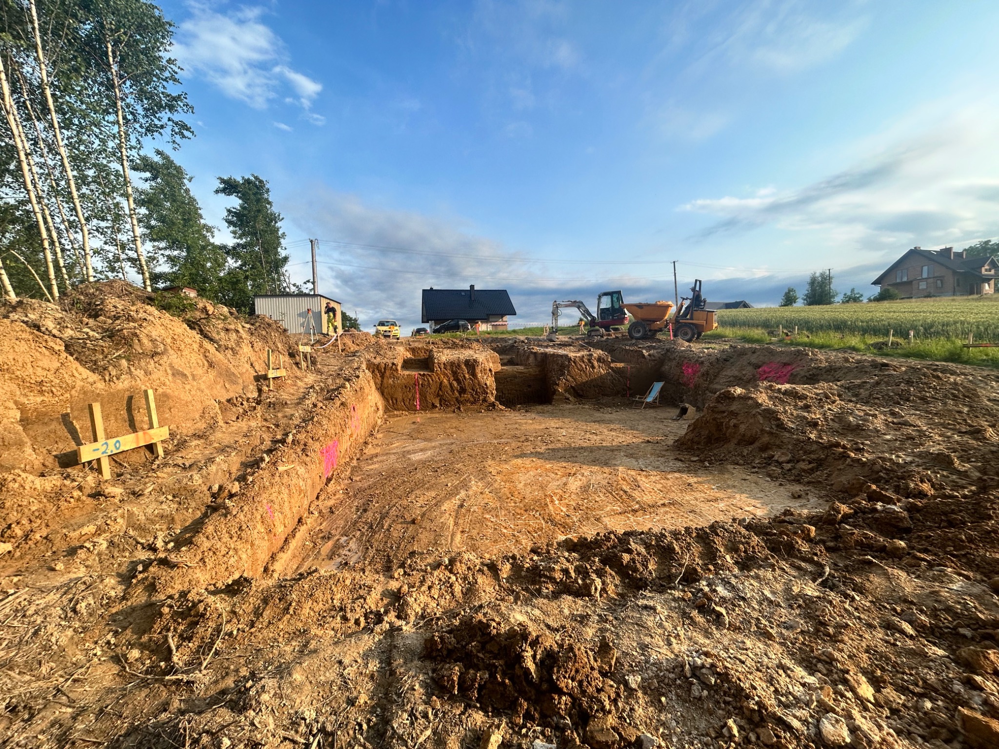 Szeroki wykop pod fundamenty budynku z widocznymi maszynami budowlanymi w tle, z zaznaczonym poziomem gruntu. Teren przygotowany pod budowę.