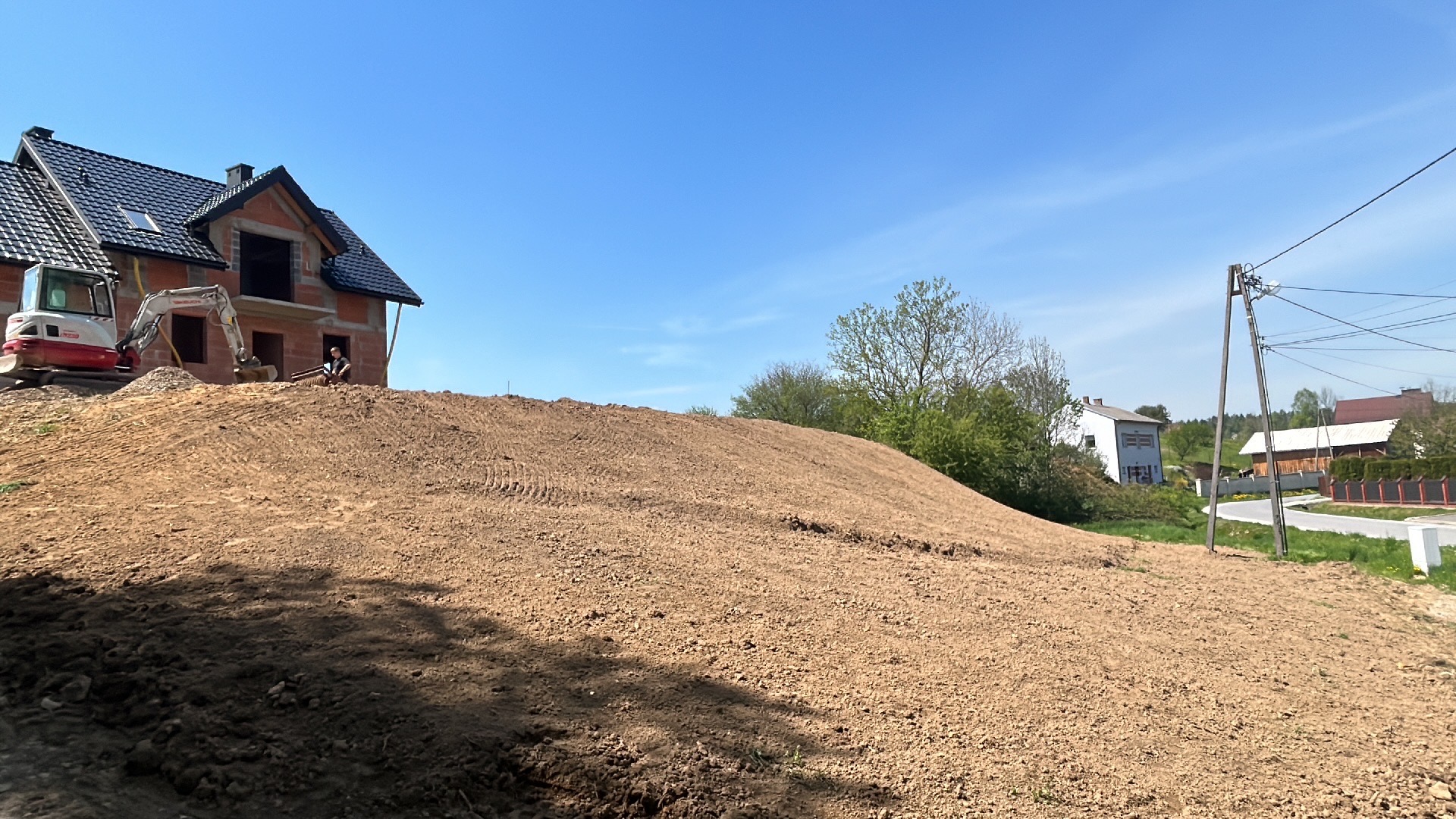Mini koparka przy domu w budowie na wzgórzu z ziemi. Ślady opon na ziemi, błękitne niebo. Prace ziemne w toku, widok na okoliczne zabudowania i słupy energetyczne.