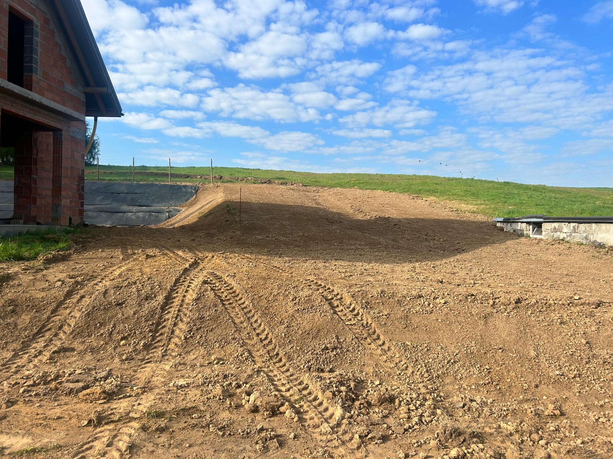 Nierówny, brązowy teren po niwelacji, z wyraźnymi śladami opon. W tle widać fragment budowy domu z czerwonej cegły i zielony pagórek pod błękitnym niebem.