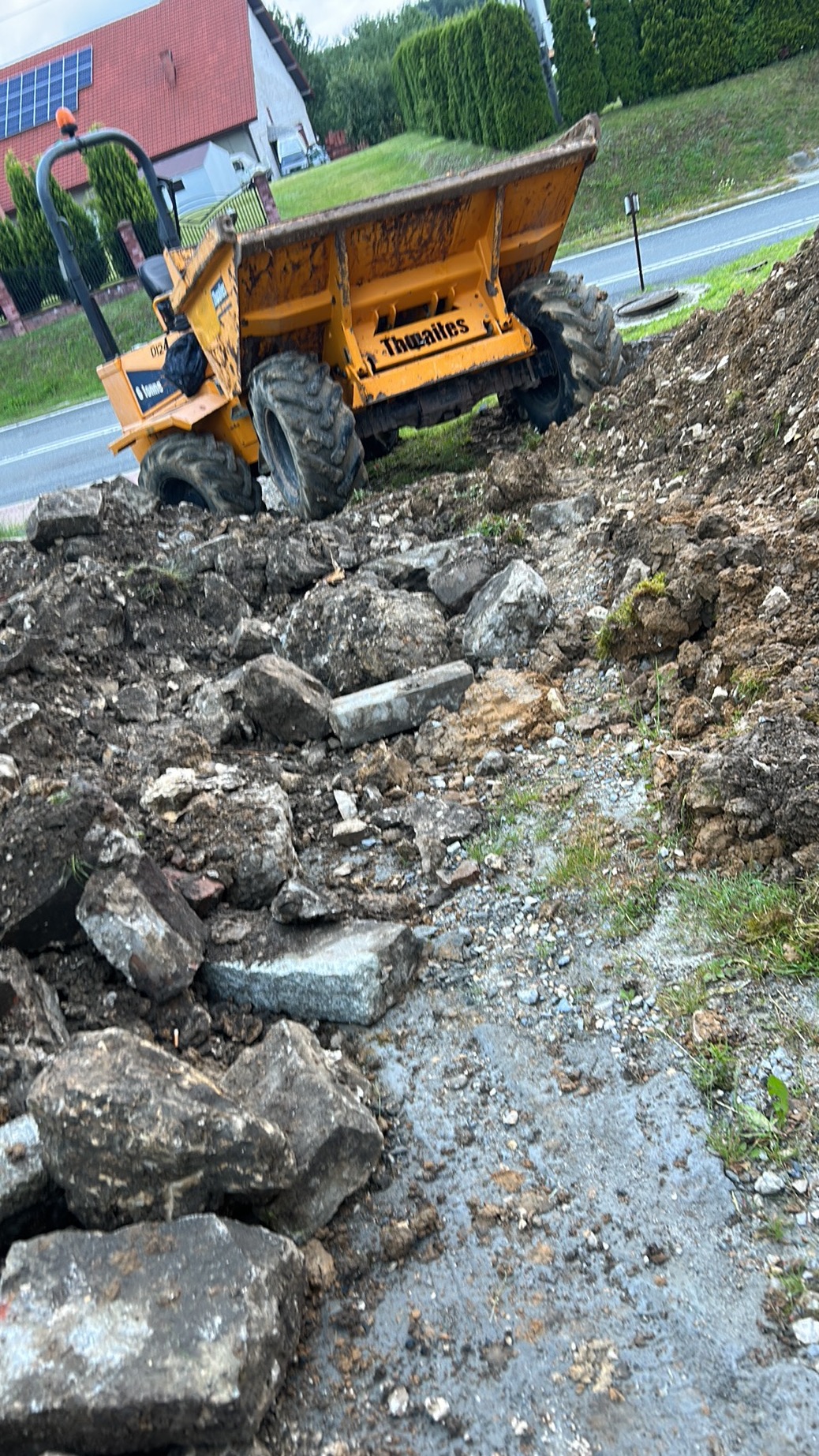 Żółta wywrotka na nierównym terenie z ziemią i kamieniami, w tle dom i zieleń. Widoczny etap prac ziemnych. Maszyna marki Thwaites.