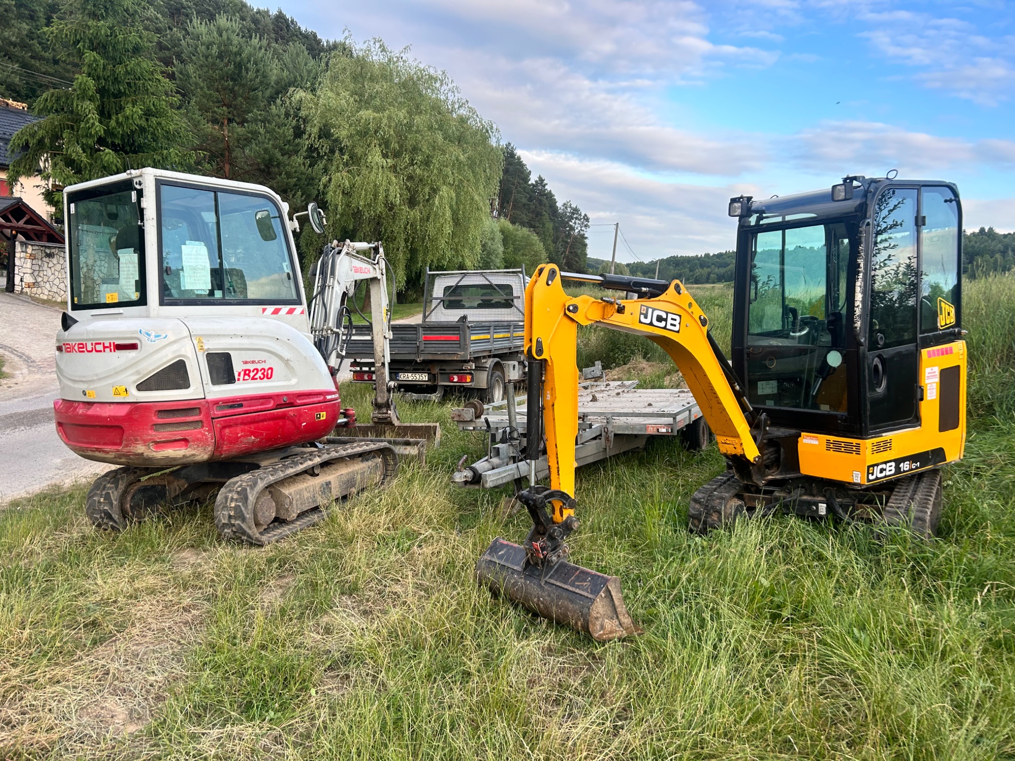 Dwie minikoparki (biało-czerwona Takeuchi TB230 i żółto-czarna JCB 16C-1) stoją na trawie obok przyczepy. W tle drzewa i fragment zabudowań. Sprzęt budowlany.