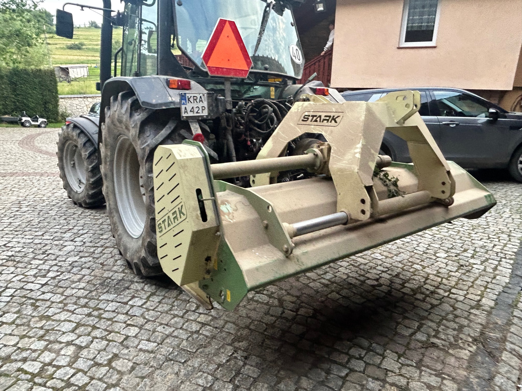 Ciężki traktor z zamontowanym frezem glebowym marki Stark, zaparkowany na brukowanej nawierzchni. Widoczny fragment budynku i samochodu w tle.