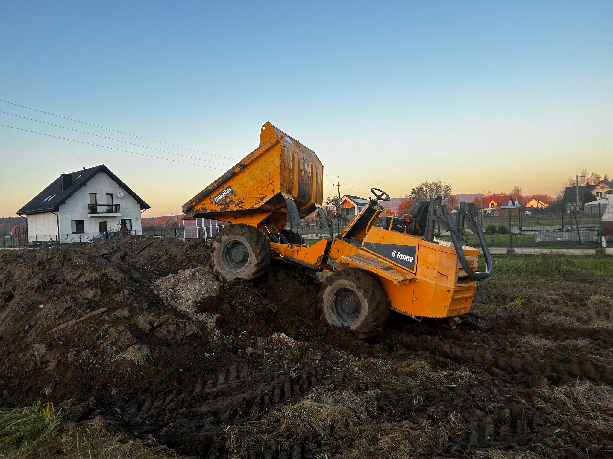 Żółta wywrotka na nierównym terenie z pryzmą ziemi, w tle dom i zachodzące słońce. Widoczne ślady opon w błocie.