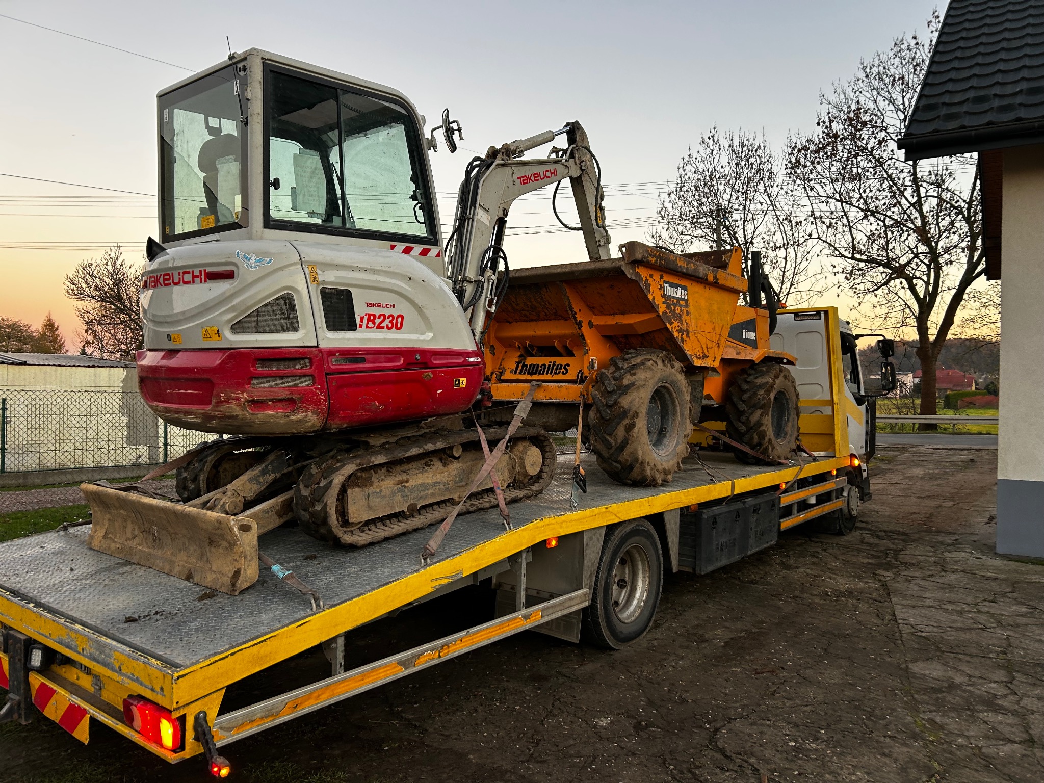 Transport minikoparki Takeuchi TB230 i wywrotki Thwaites na niskopodwoziowej lawecie w jesienny dzień. Sprzęt budowlany zabezpieczony pasami transportowymi.