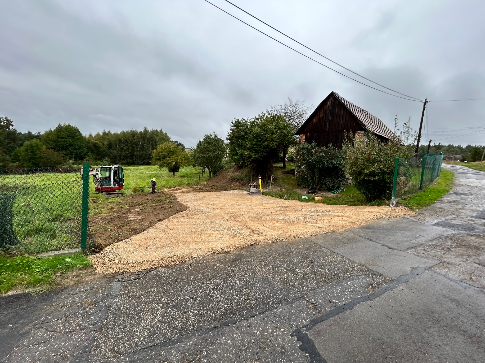 Przygotowany teren pod budowę z użyciem minikoparki, widoczny operator w odblaskowej kamizelce. W tle drewniany budynek i zieleń. Pochmurny dzień.