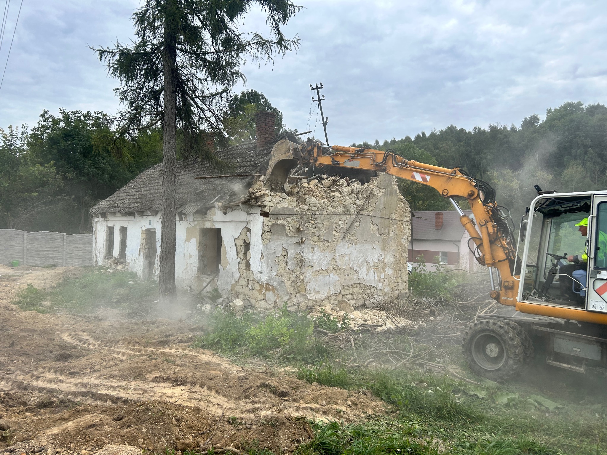 Żółta koparka kołowa w trakcie rozbiórki murowanego, parterowego budynku z ceglanym kominem. W tle drzewa i fragment betonowego ogrodzenia.